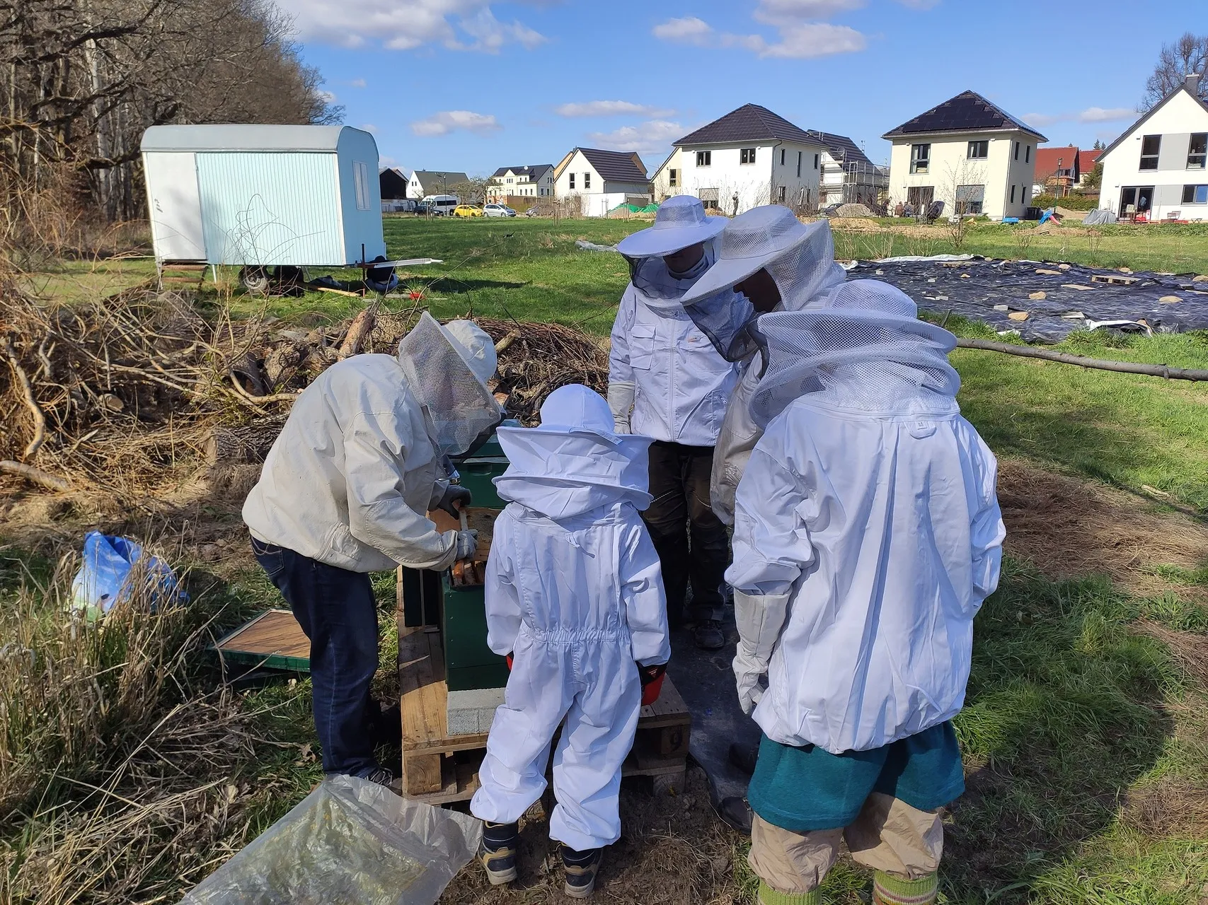 Kinder in weißen Imkeranzügen stehen mit einer erwachsenen Imkerin am geöffneten Bienenstock